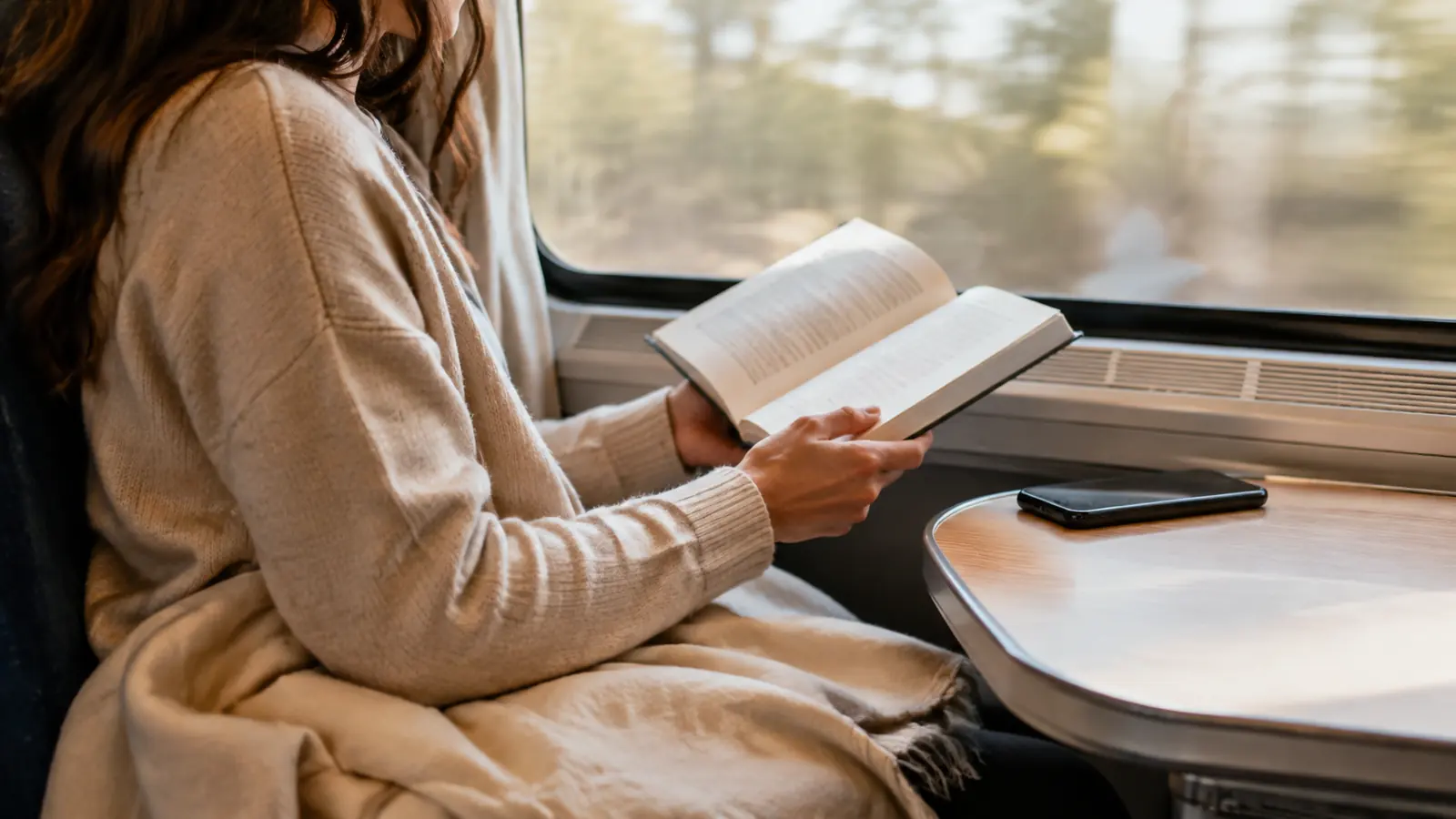 A reader holding an open Bible near a train window with a phone resting face down beside a bookmark